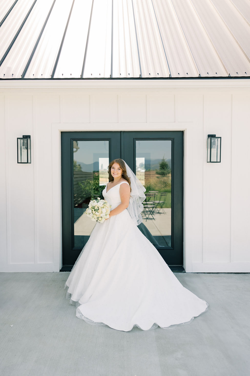 A bride stands on a porch smiling with her white bouquet