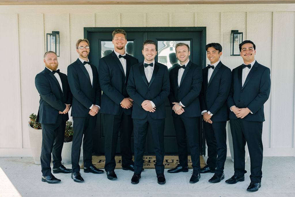 A groom stands on a porch with his groomsmen all in black