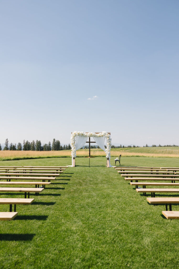 A view down the aisle of a set up farmhouse on green bluff outdoor wedding ceremony with flower covered arbor and wooden cross