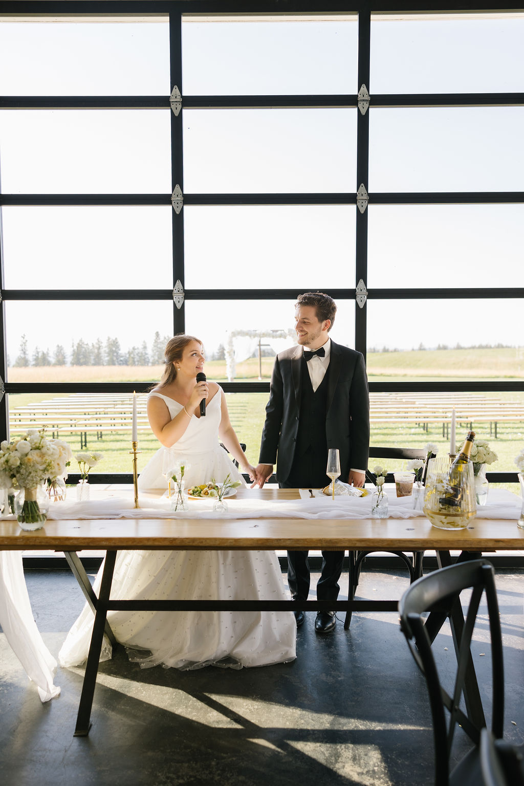 A bride gives a toast while smiling at her groom at their wooden head table during their farmhouse on green bluff reception