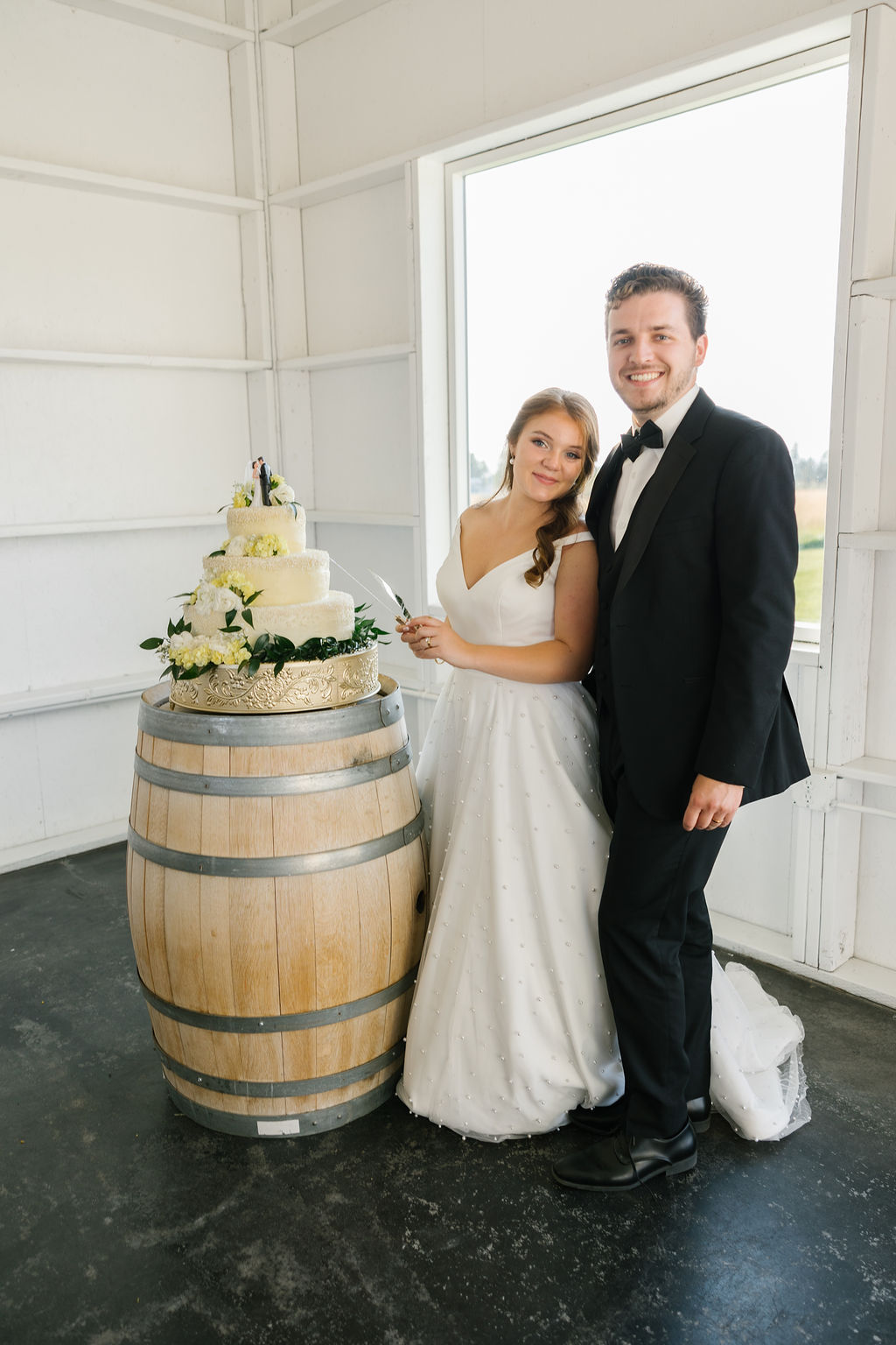 A bride and groom smile while preparing to cut the three tier cake on a barrel