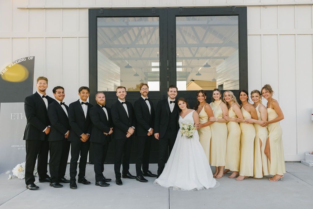 A bride and groom stand by the entrance to farmhouse on green bluff with their large wedding party in black and gold