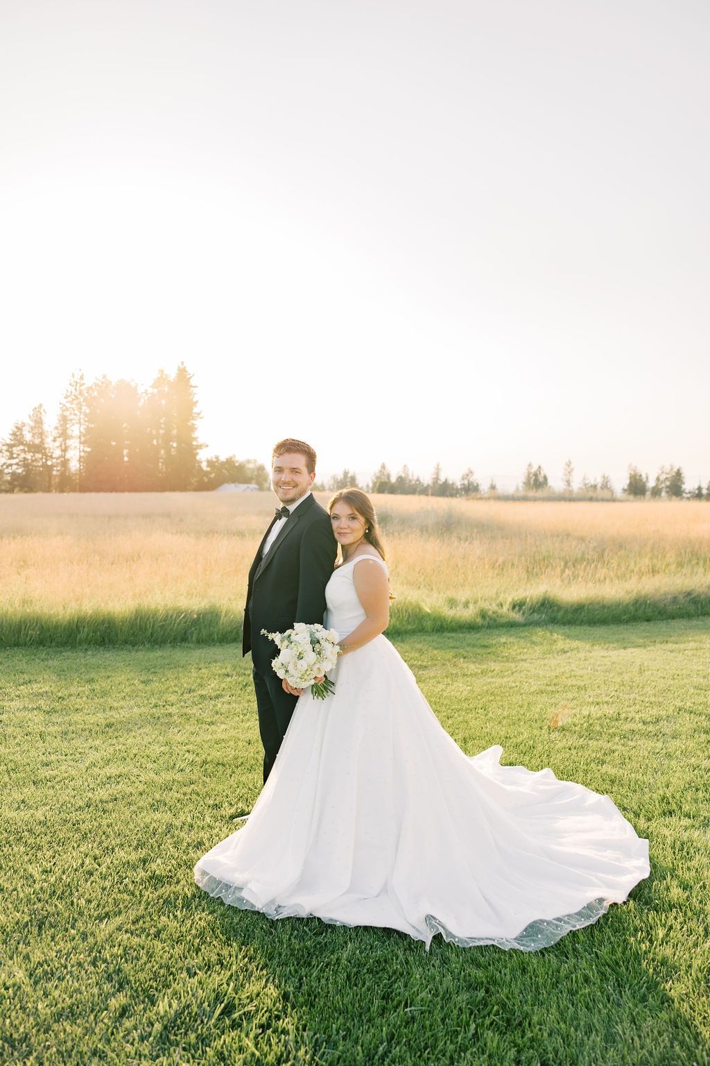 A bride leans on the shoulder of her groom as they stand in a pasture at sunset