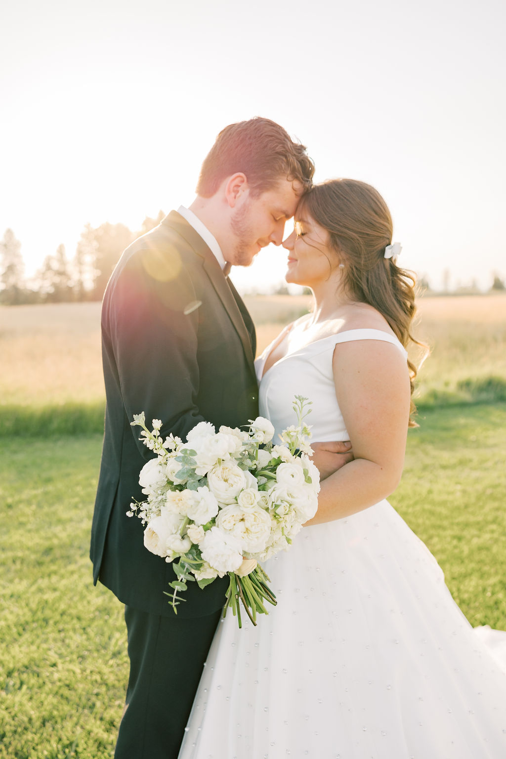 Newlyweds touch foreheads and snuggle while standing in a pasture at sunset at farmhouse on green bluff