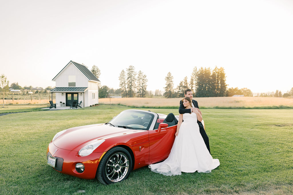 A bride and groom lean against a red convertable in the lawn of farmhouse on greenbluff