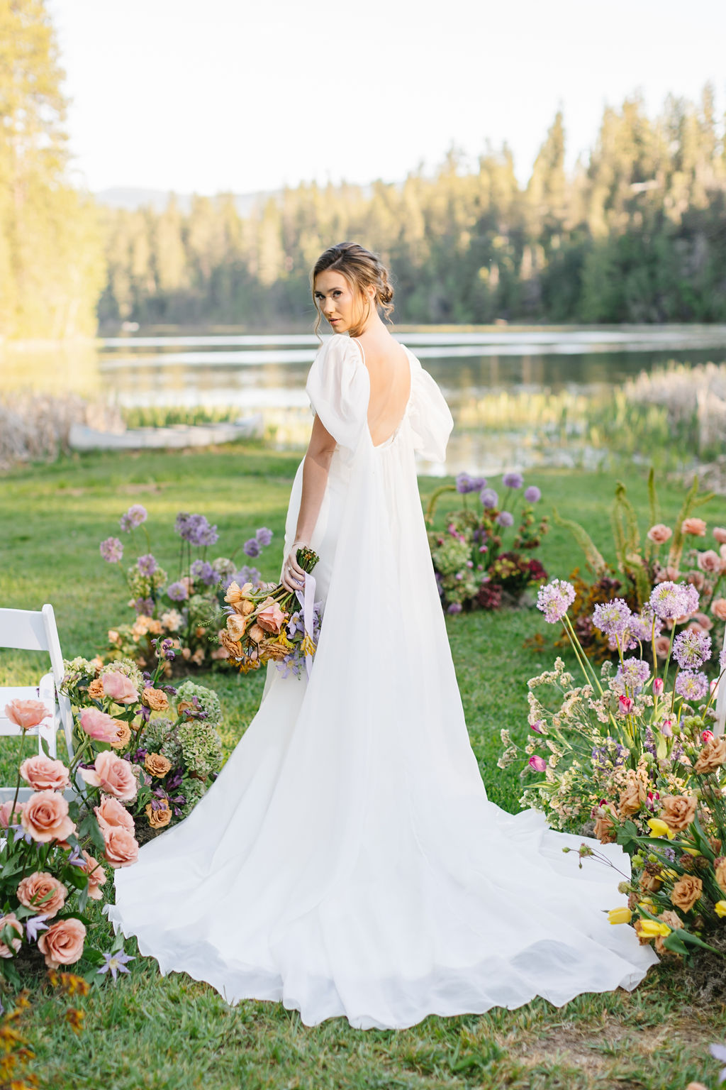 A bride looks over her shoulder while standing in the aisle of her ceremony location with colorful flowers and white chair at lakefront wedding venues coeur d'alene