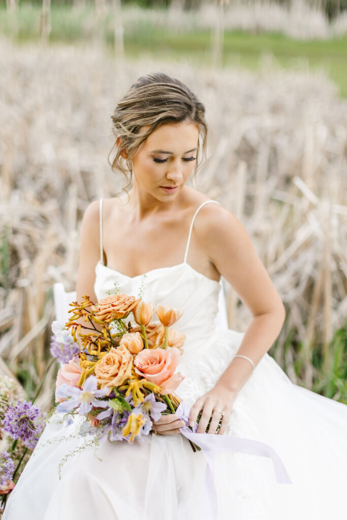 A bride gazes down her shoulder while sitting by some tall grasses by a lake