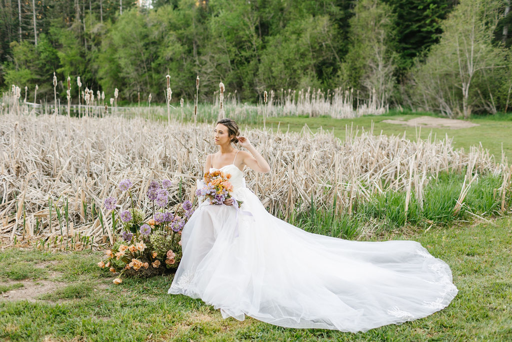 A bride sits in a lawn fixing her hair with a long train holding her orange bouquet