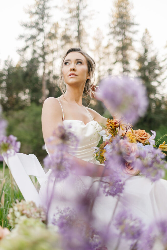 A bride sits in a white chair in a lawn holding her flowers at sunset