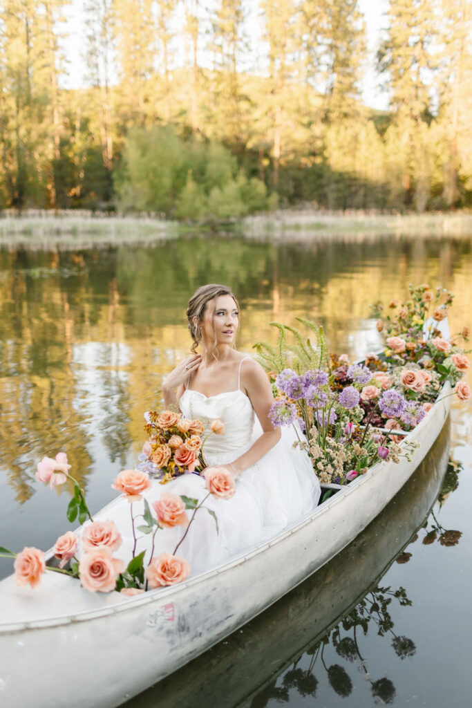 A bride looks over her shoulder while sitting in a canoe filled with colorful flowers on a calm day