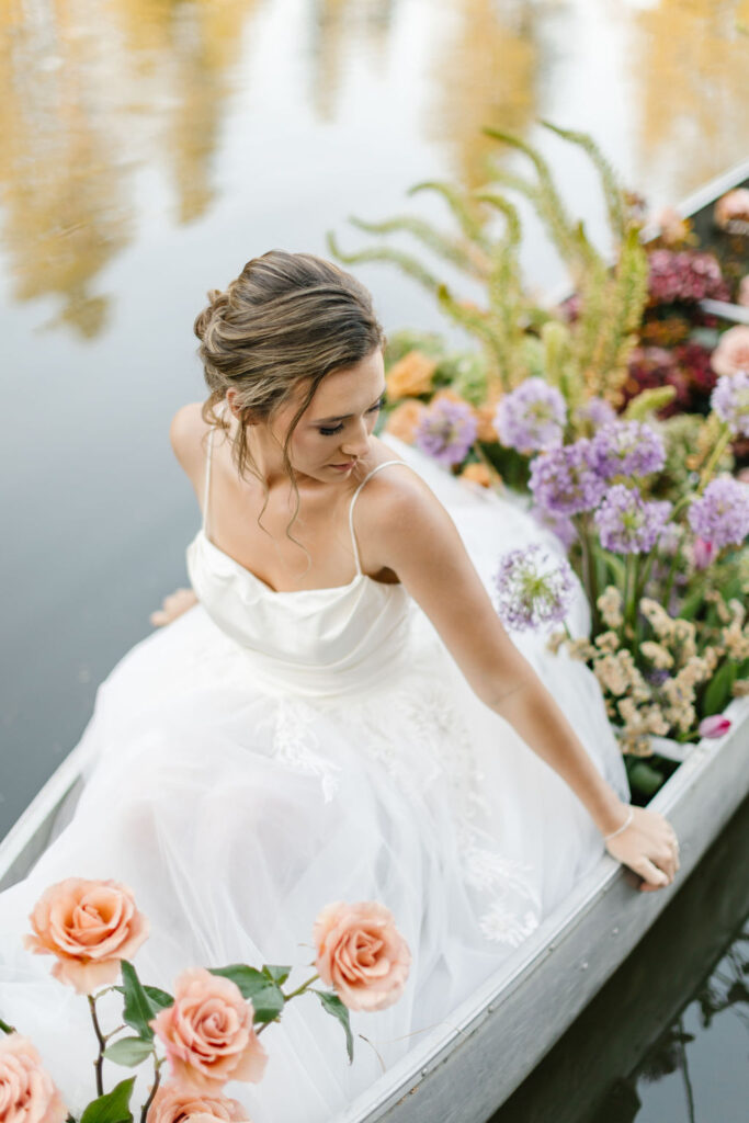 A bride sits in her dress in a canoe with colorful flowers while floating in a lake