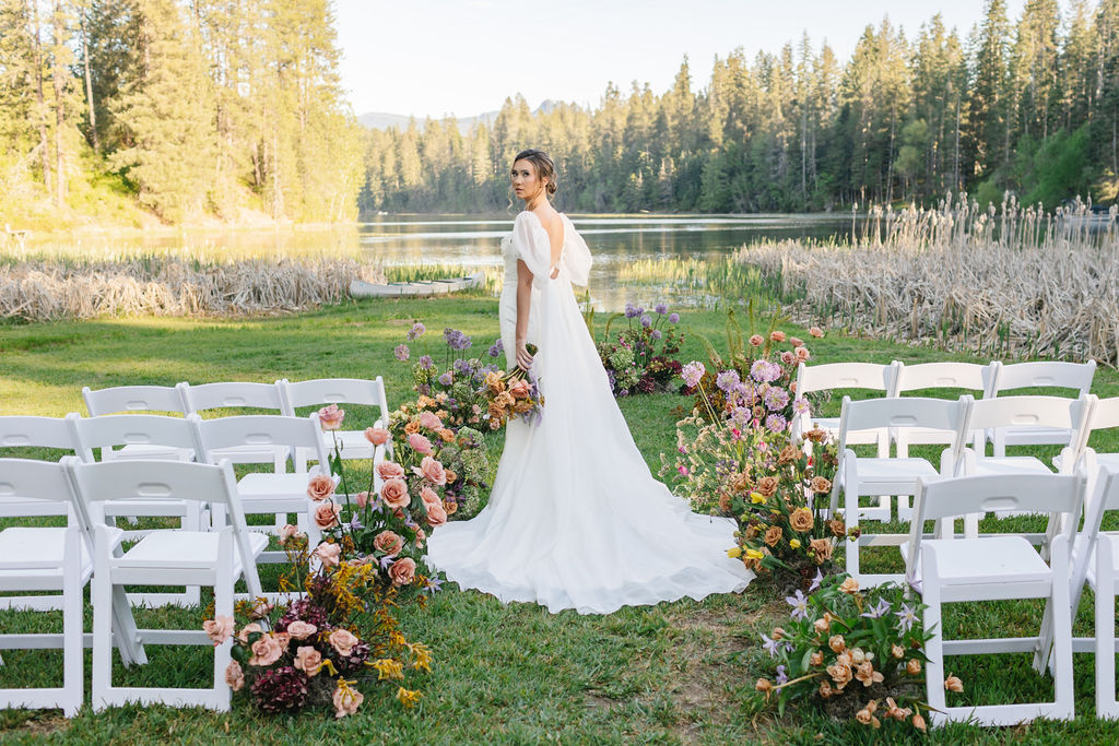 A bride walks down the empty aisle on a lake of her ceremony location lined with colorful flowers