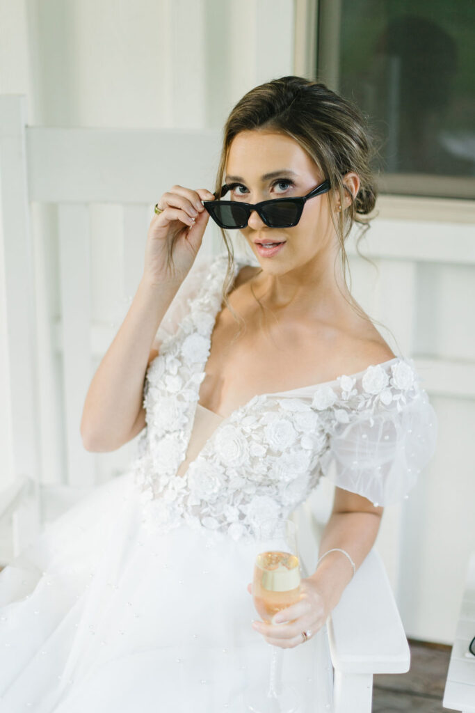 A bride peeks over her sunglasses while sitting on a porch with a glass of champagne