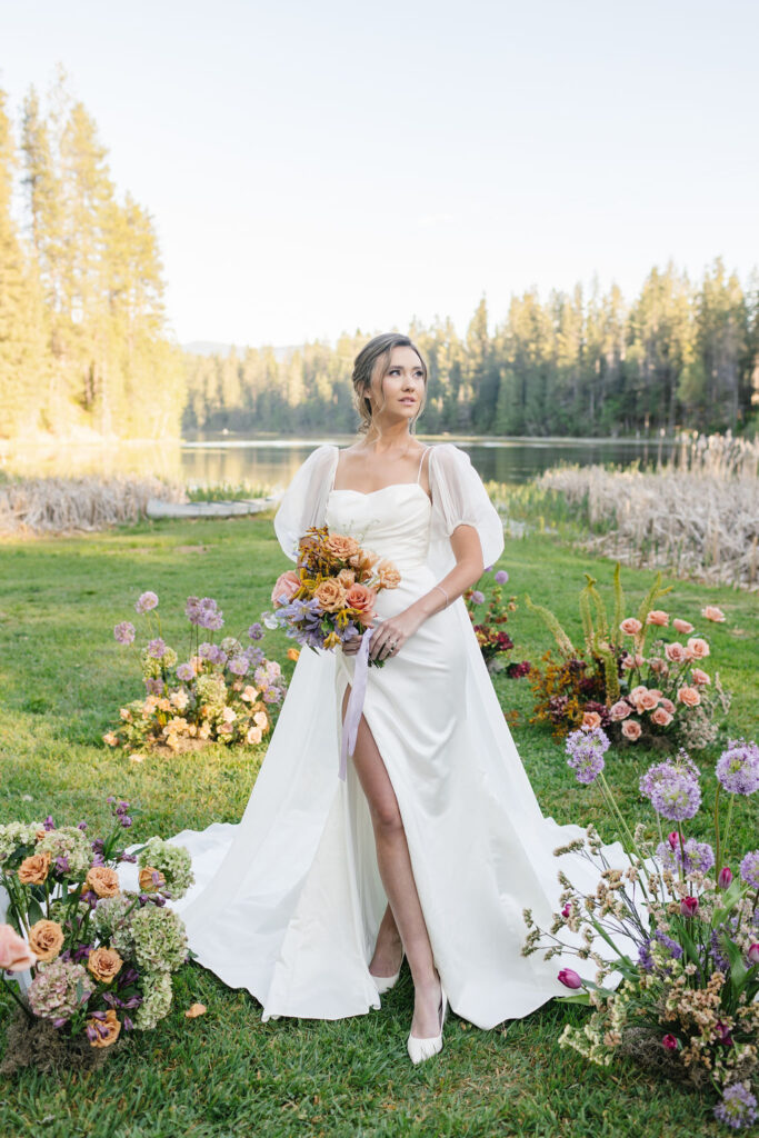 A b ride smiles over her shoulder while walking among multiple colorful flower arraignments on the ground at lakefront wedding venues coeur d'alene