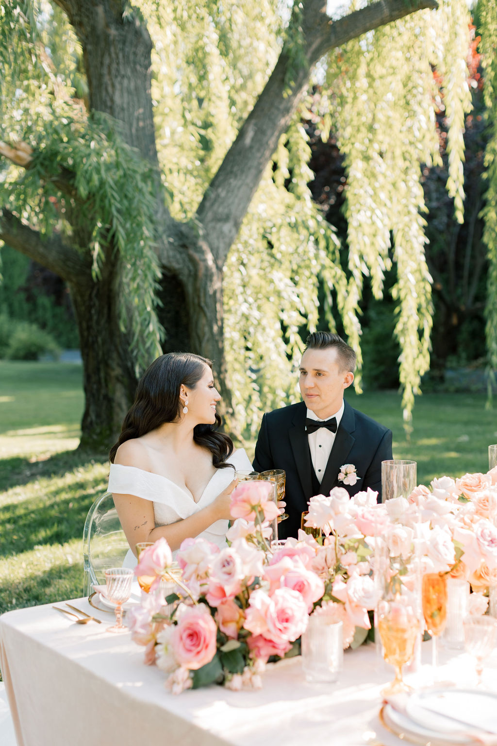 A bride and groom laugh and toast at their head table under an old willow tree