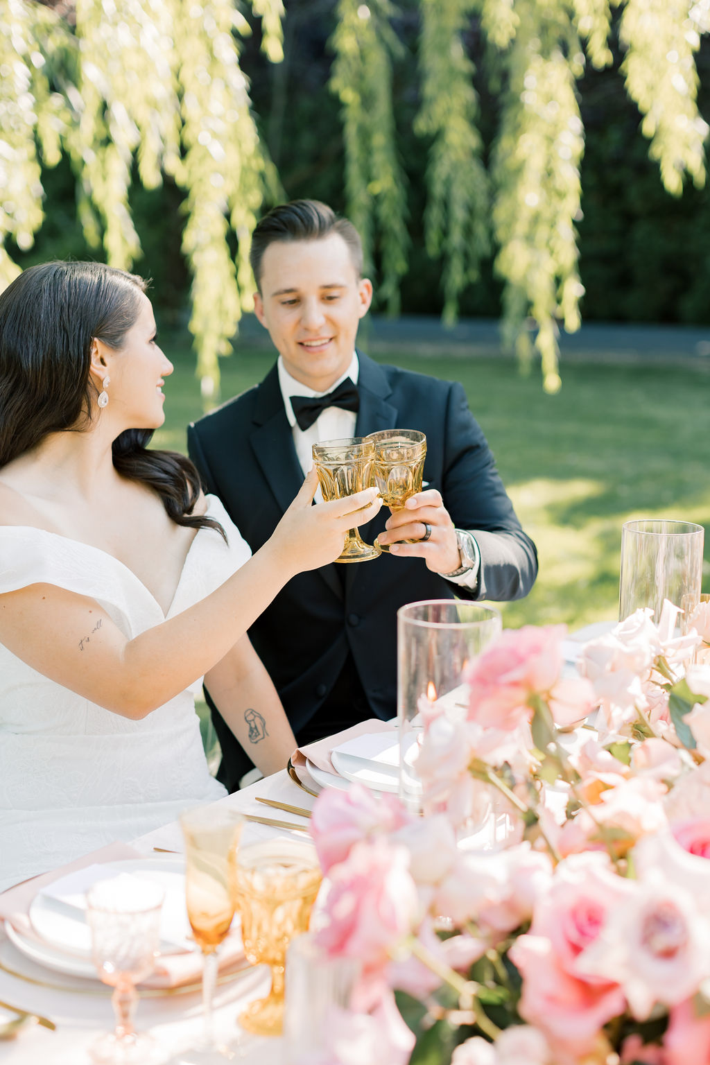 A bride and groom toast at their head table covered in pink roses under a willow at sunset at promise garden pasco