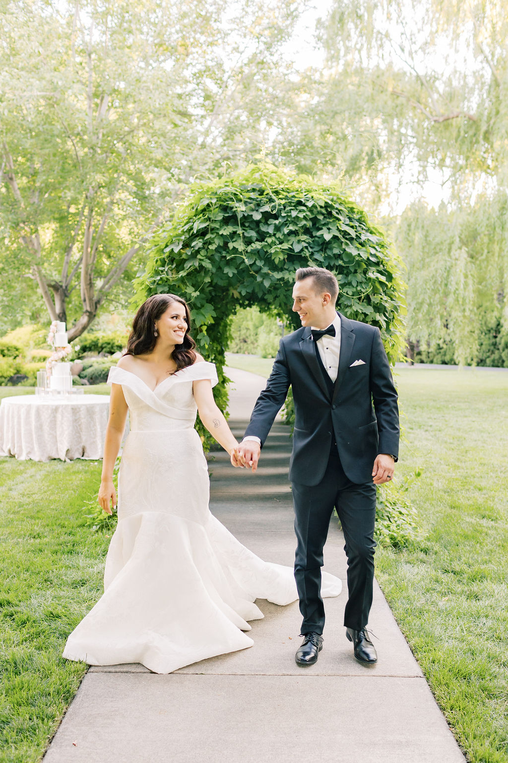 A bride and groom hold hands and walk up a garden sidewalk smiling at each other during their promise garden pasco wedding