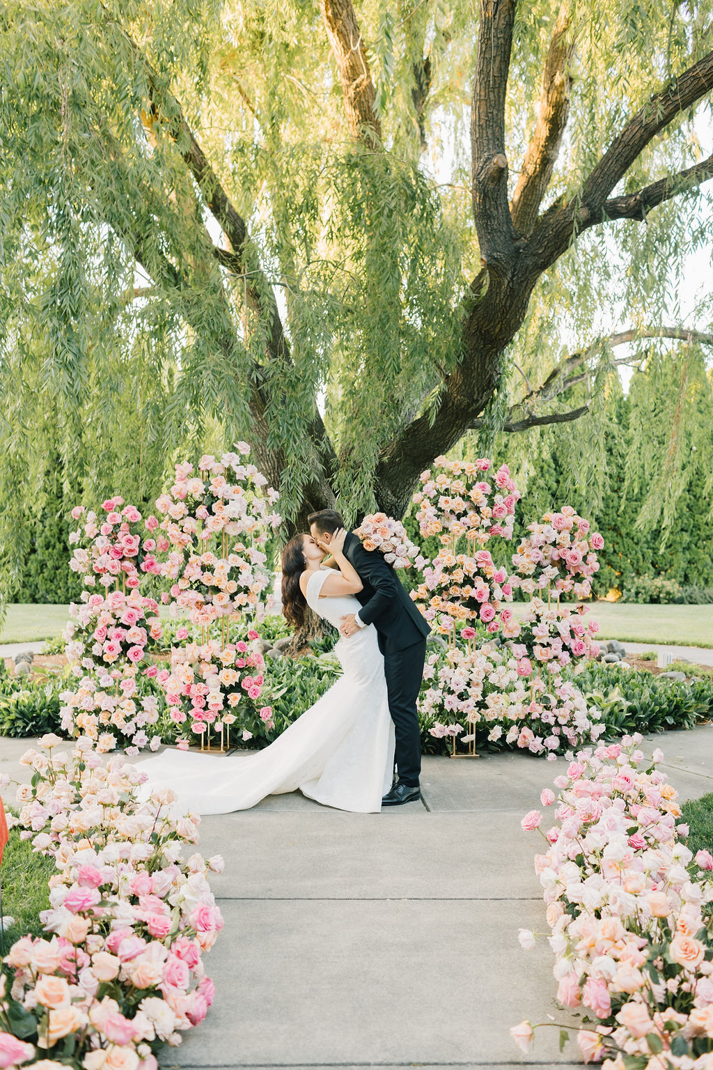 Newlyweds kiss under a giant willow tree surrounded by white and pink roses to finish their promise garden pasco wedding ceremony