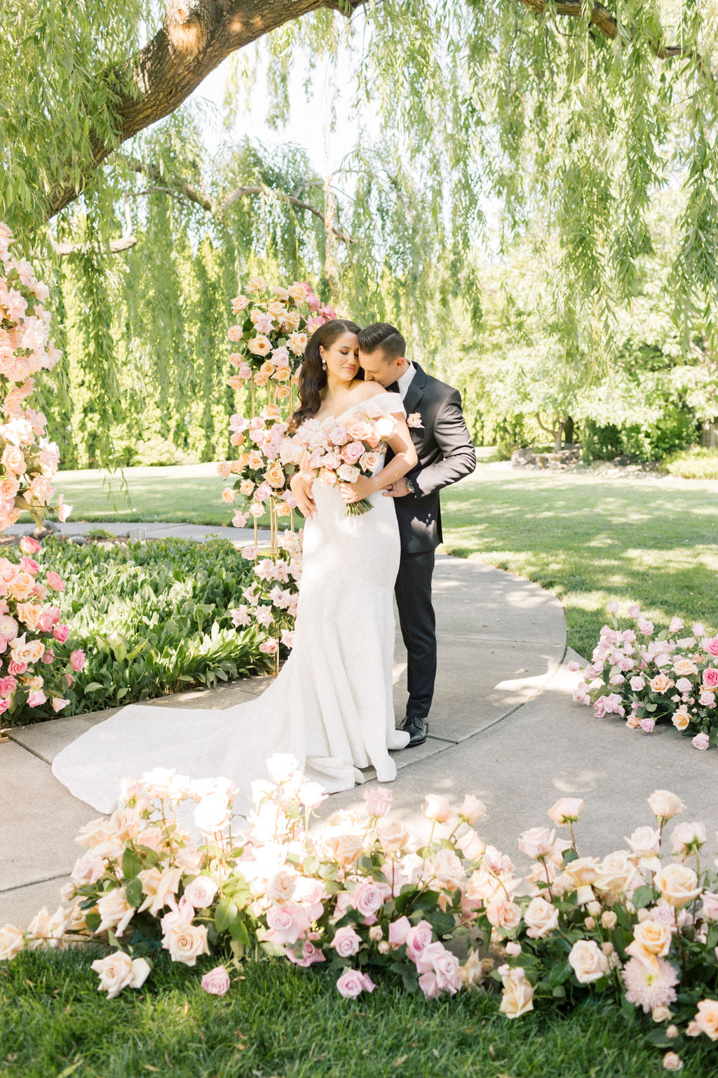 A groom kisses the shoulder of his smiling bride under a willow tree surrounded by roses