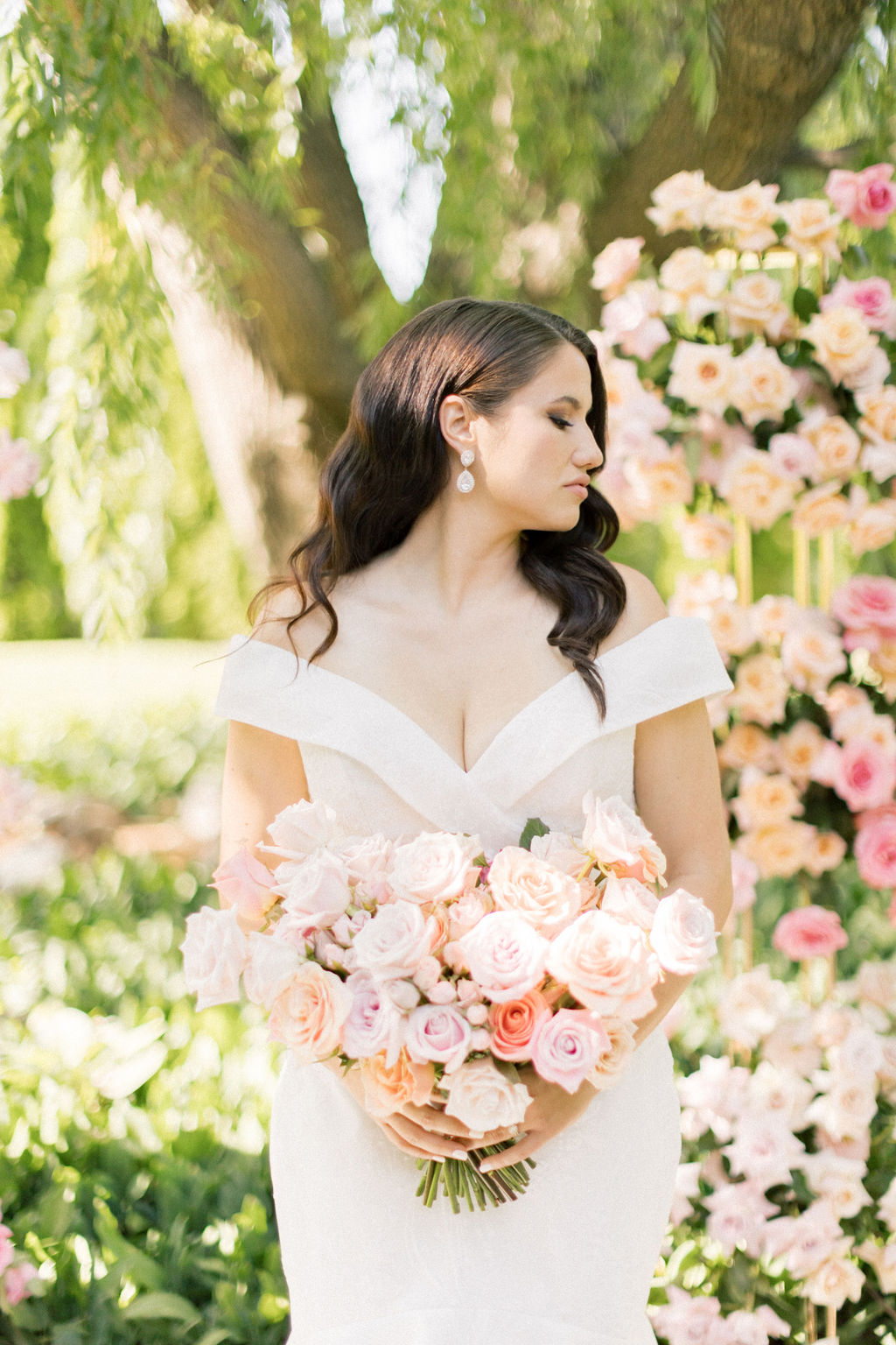 A bride looks over her shoulder while holding her bouquet in a pink rose garden