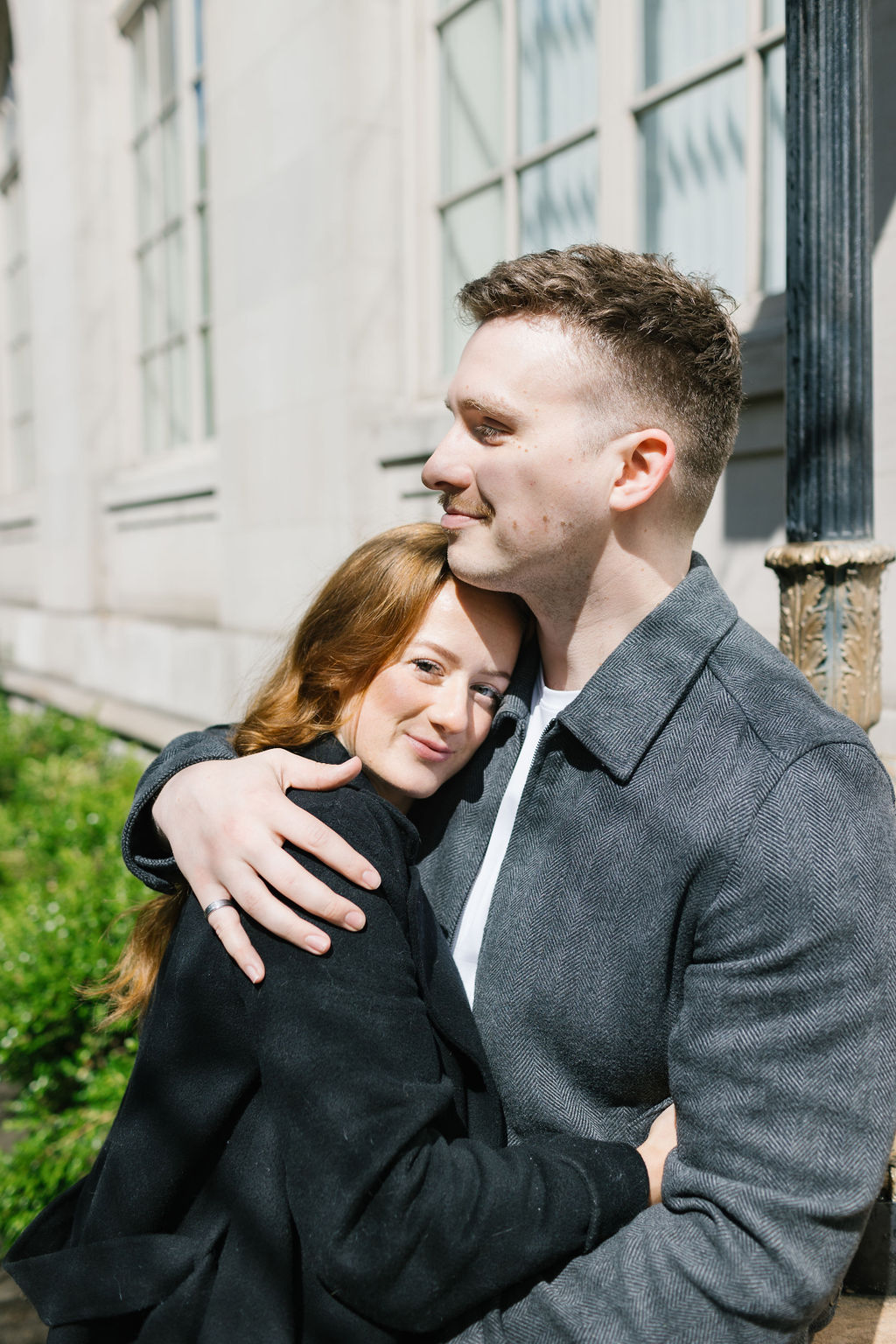 A couple snuggles against an ornate building in jackets in the sun