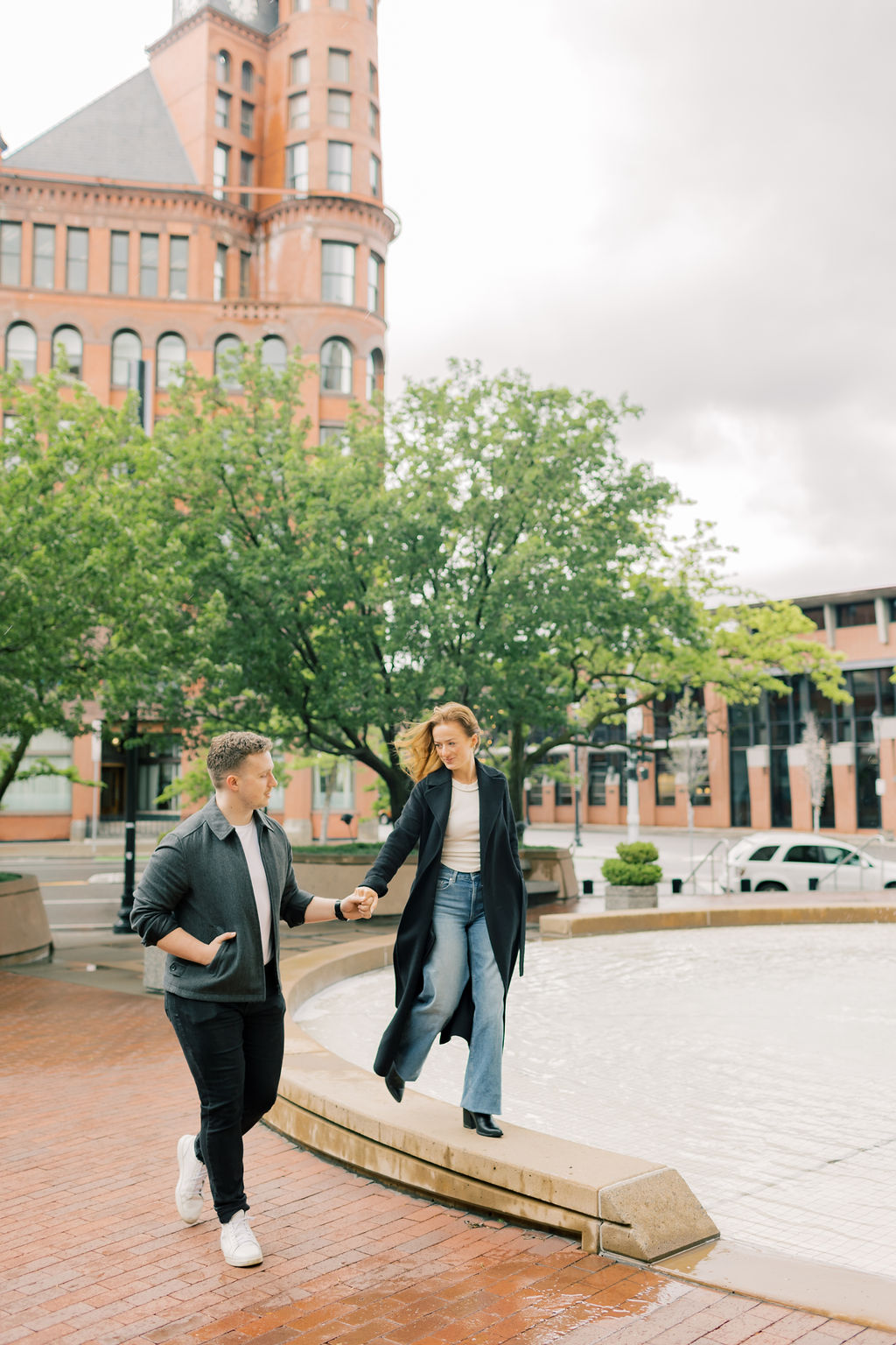 A woman walks on a fountain ledge while holding hands with her fiancee