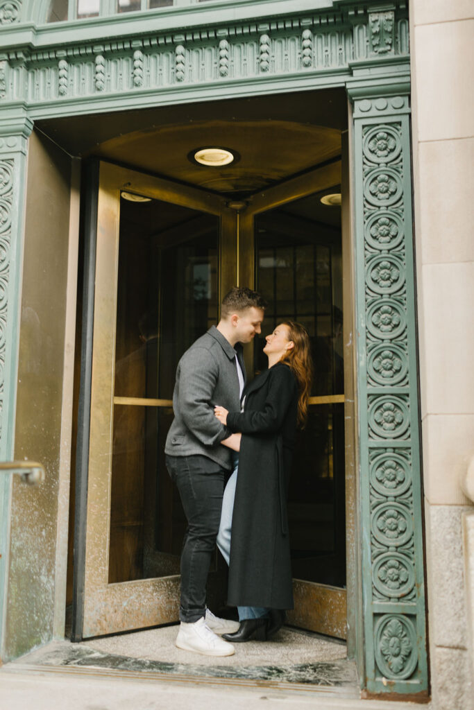 A couple hugs while in a revolving door entrance with ornate trim