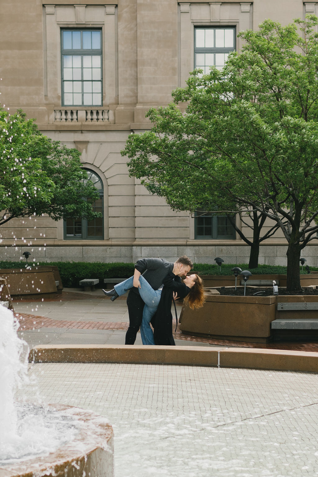 Newly engaged couple dips and kisses by a park fountain in downtown