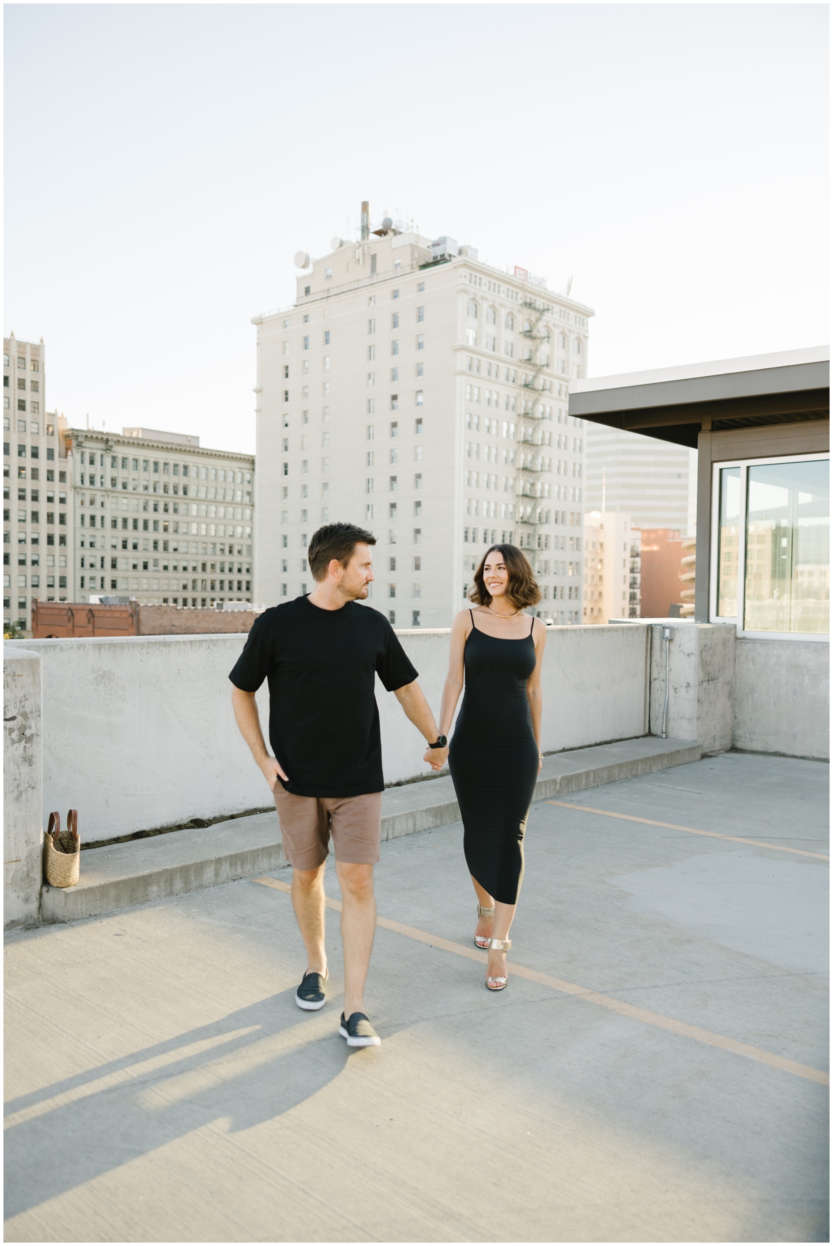 A newly engaged couple walks hand in hand on a rooftop parking garage in a downtown metro