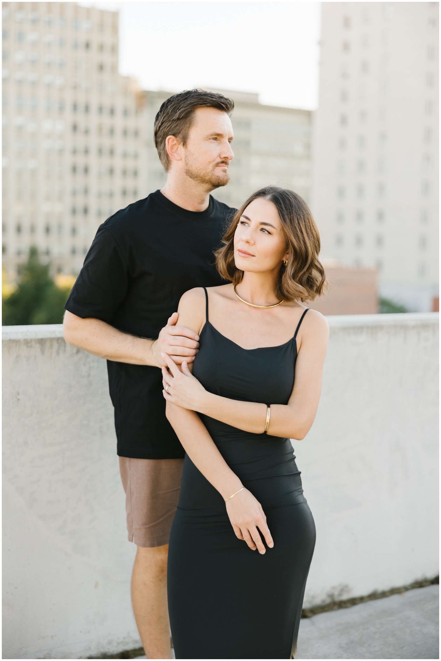 A woman in a black dress leans against her fiancee on a rooftop during their rooftop engagement photos