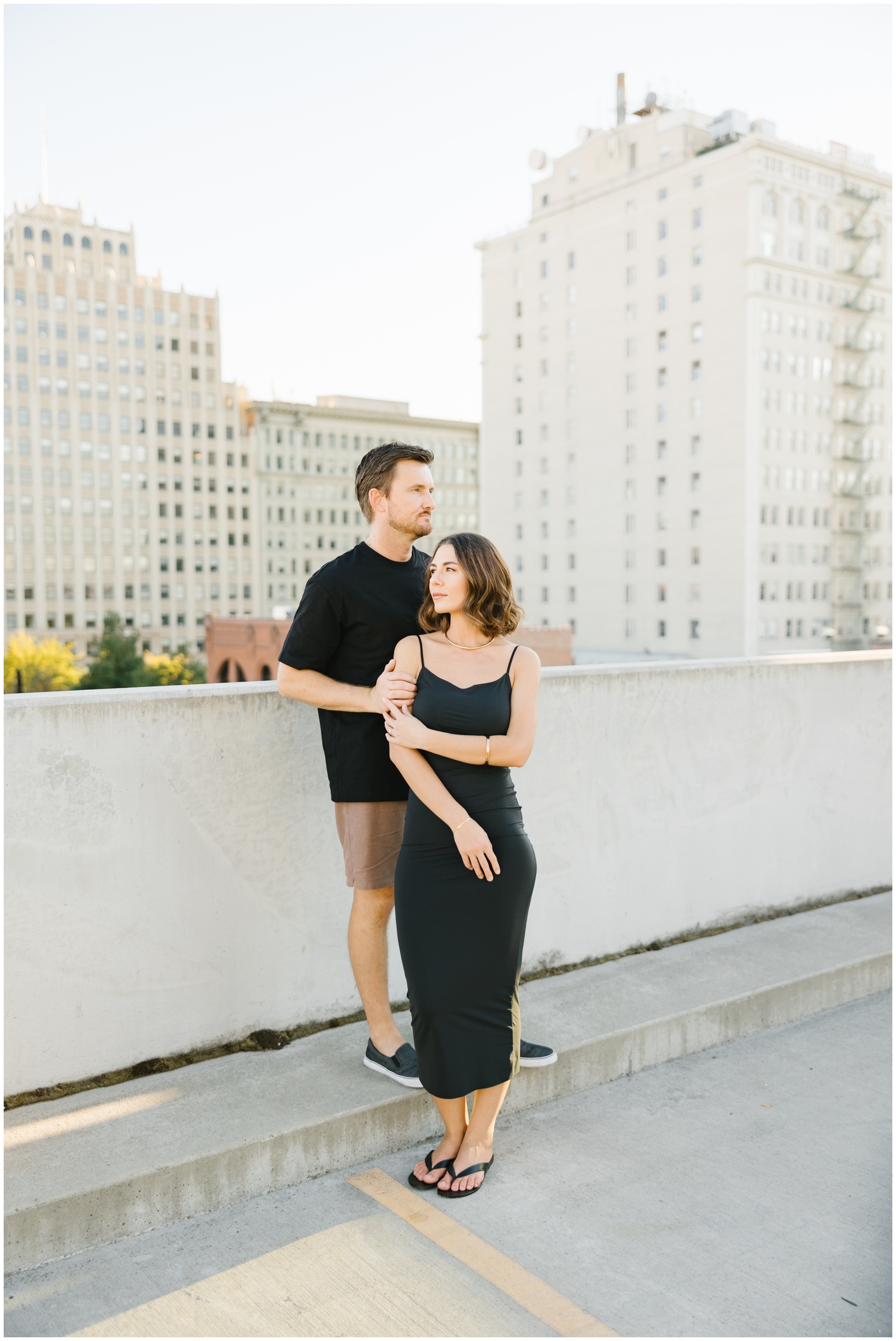 An engaged couple in black stand by a wall in downtown during their rooftop engagement photos