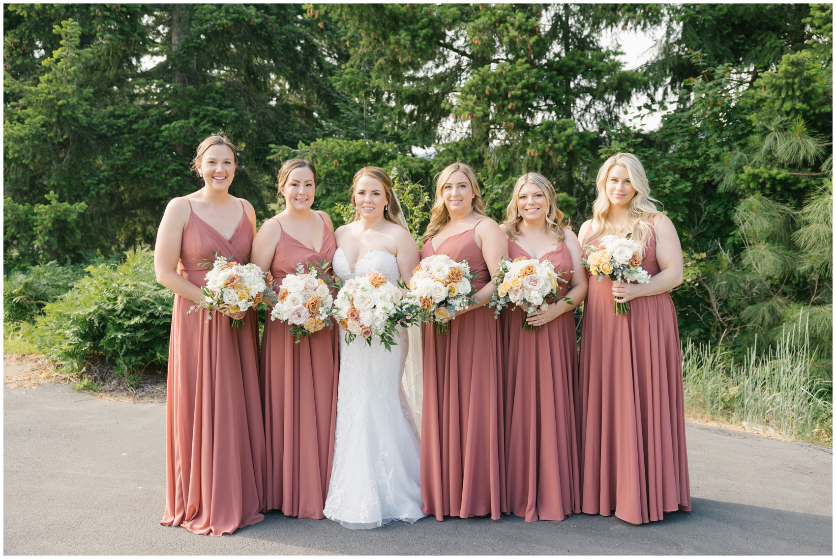 A bride stands with her bridesmaids in pink dresses while holding their bouquets on a road by a forest