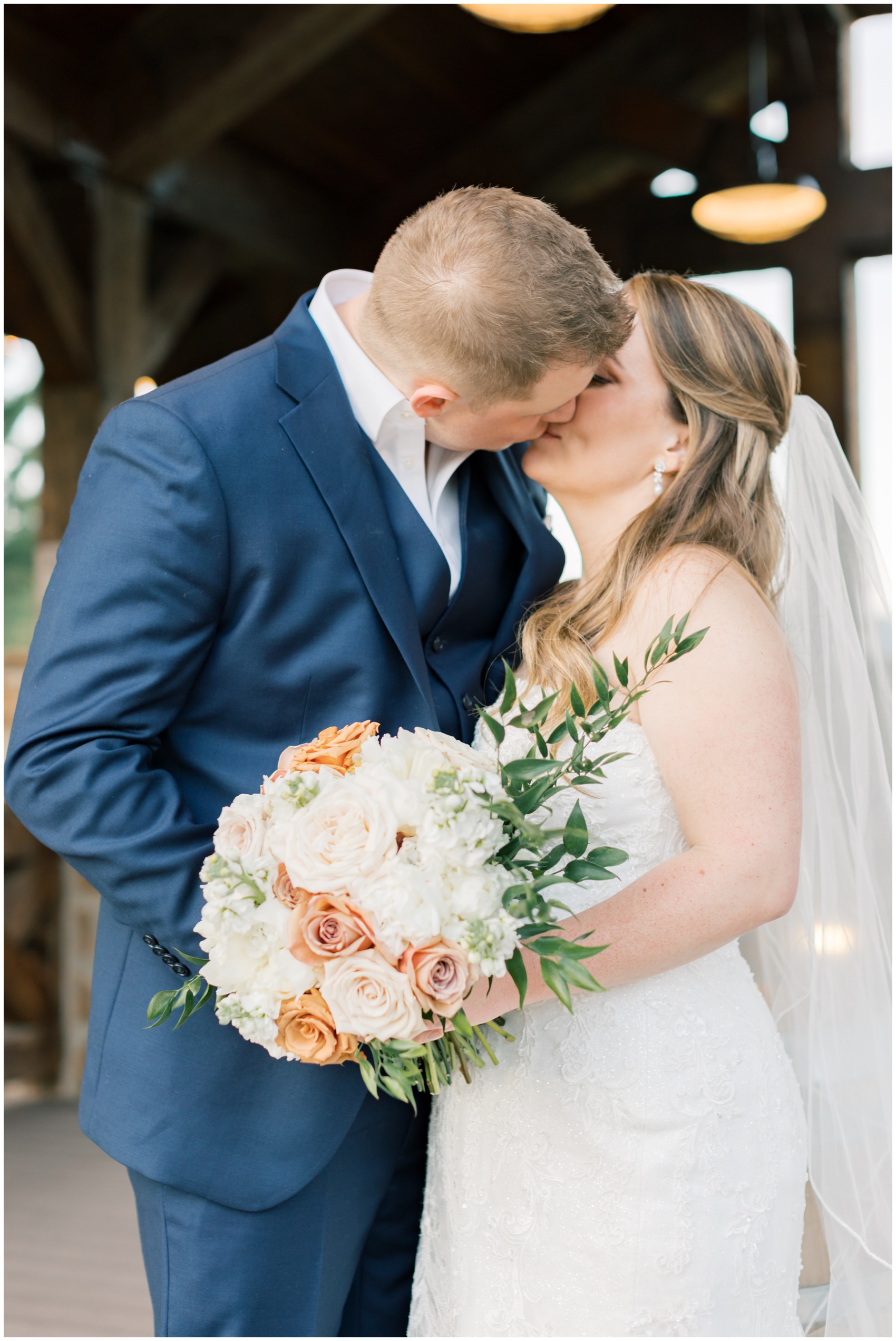 Newlyweds kiss on a patio holding the pink and white bouquet with groom in a blue suit