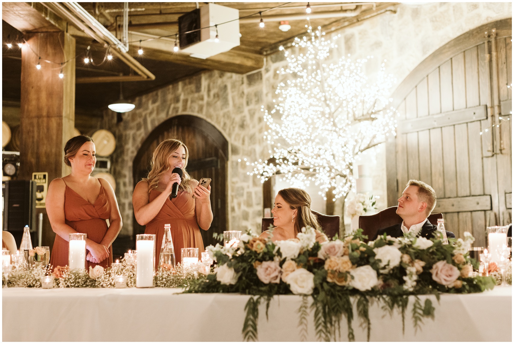 A bride and groom smile while listening to bridesmaids speech at their head table covered in flowers