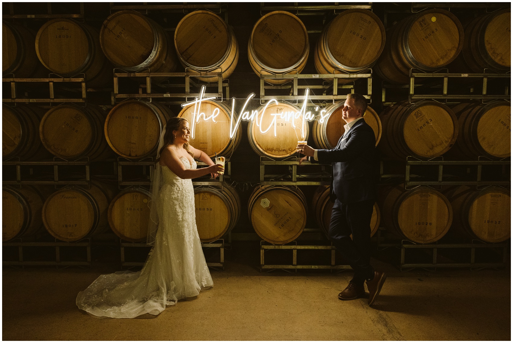 Newlyweds lean on a wall of wine barrels with a custom neon sign and drinks