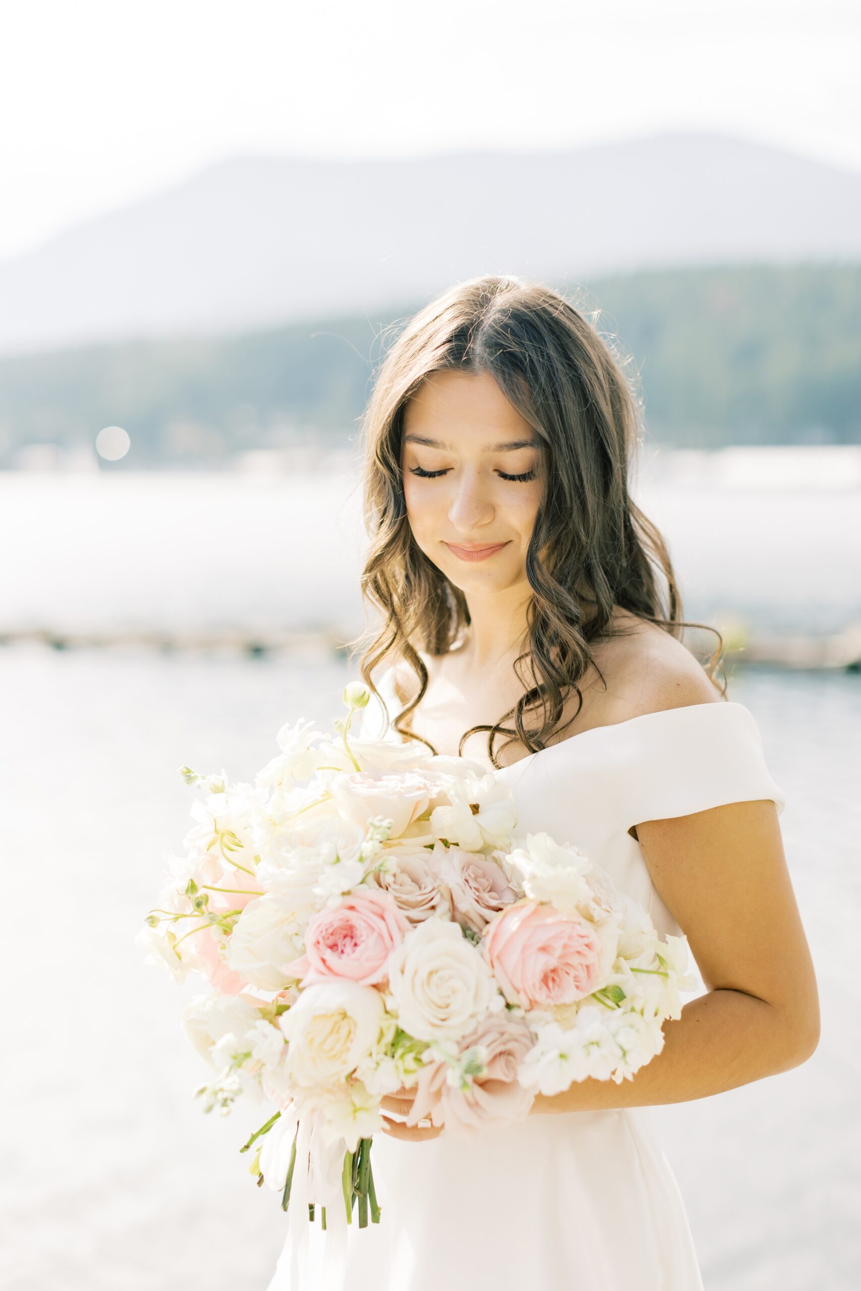 A bride stands by a lake smiling down at her white and pink bouquet