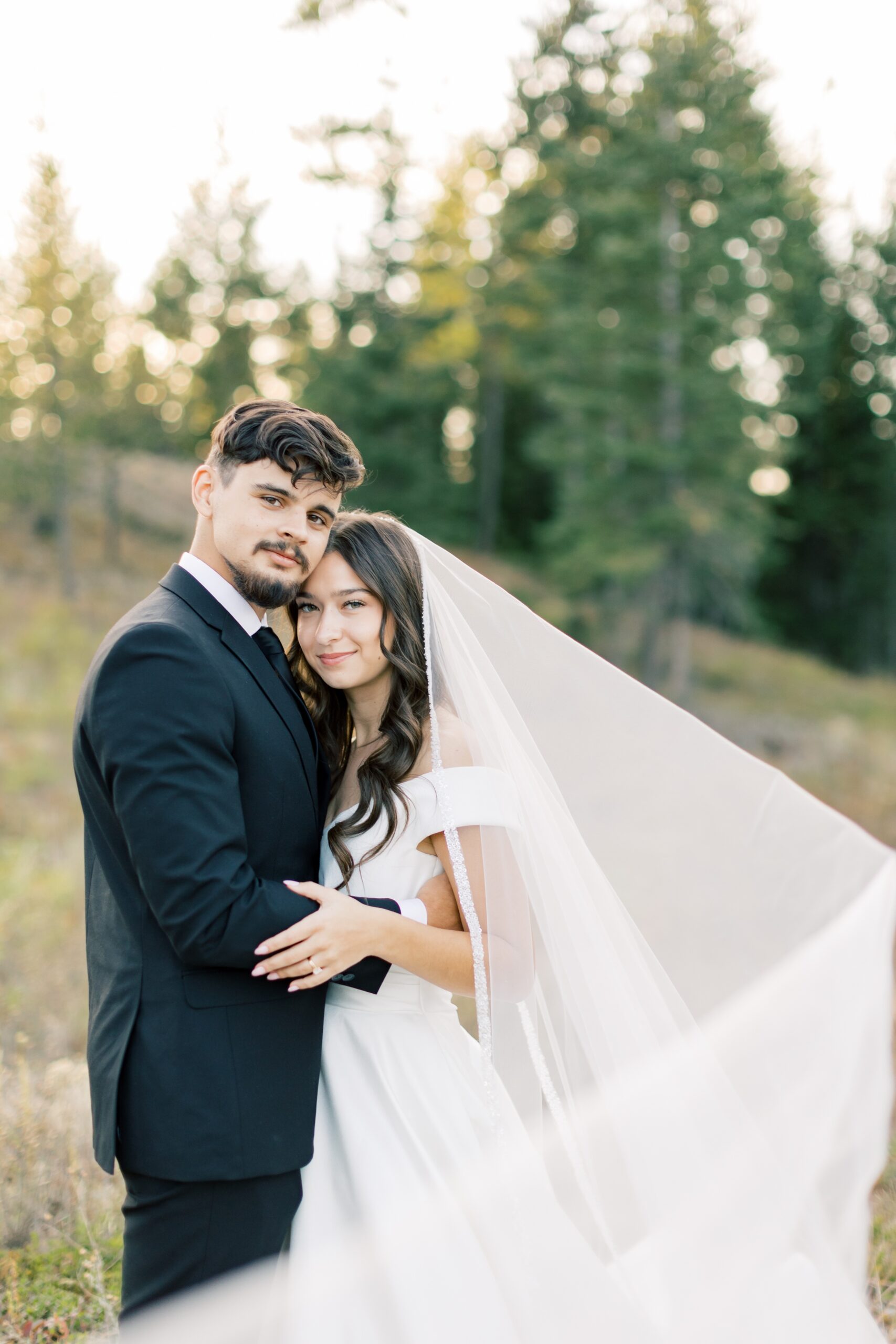 A happy couple snuggles on the side of a hill at sunset with the veil flowing around them at one of the Hayden Lake wedding venues