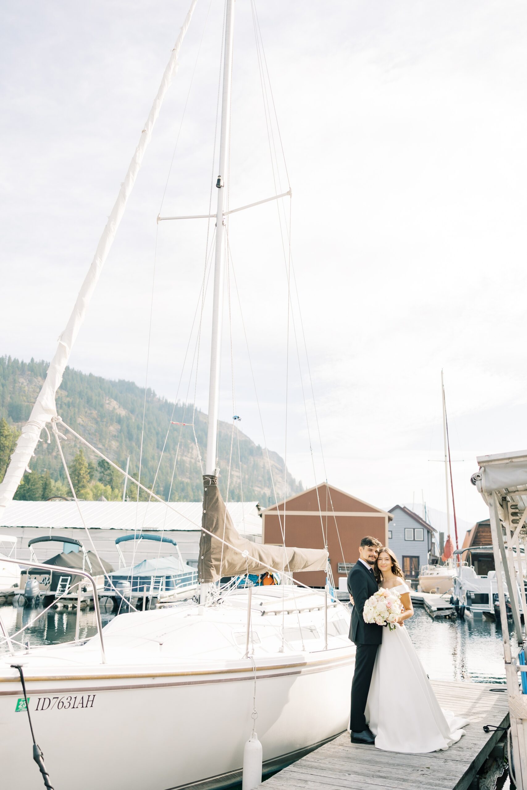 A happy couple snuggles while standing on a dock by a sailboat in a marina at one of the Hayden Lake wedding venues