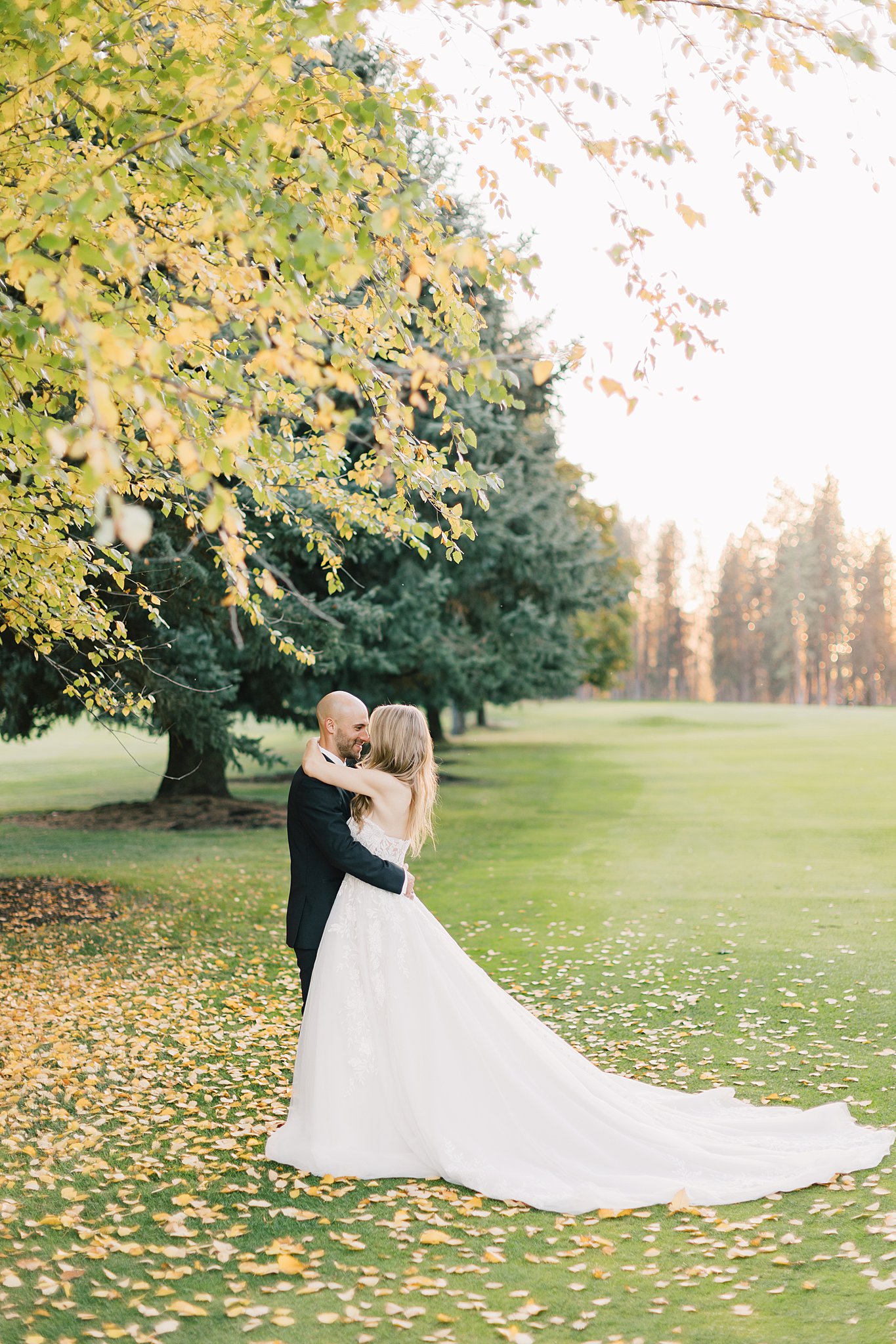 A bride and groom embrace under a yellowing tree in a fairway at sunset