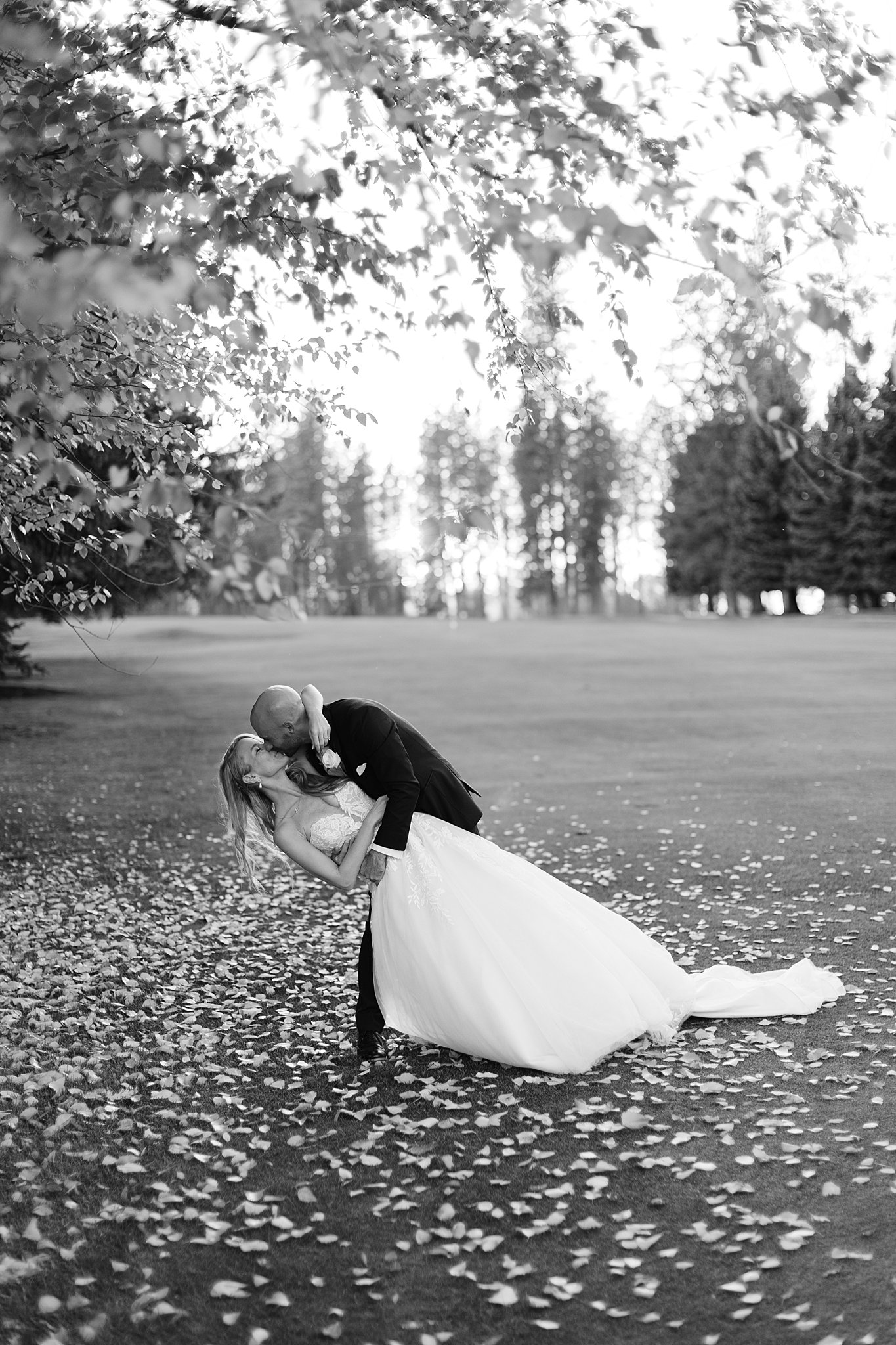 A groom dips and kisses his bride under a tree in a field covered in fallen leaves at their Kalispel Country Club Wedding