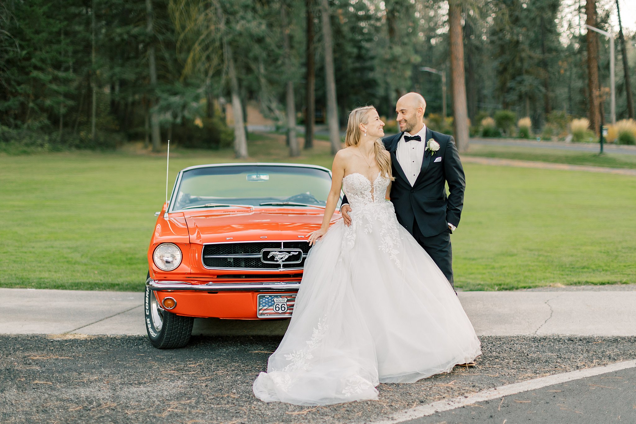 A bride and groom smile at each other as they lean on a 1966 orange ford mustang