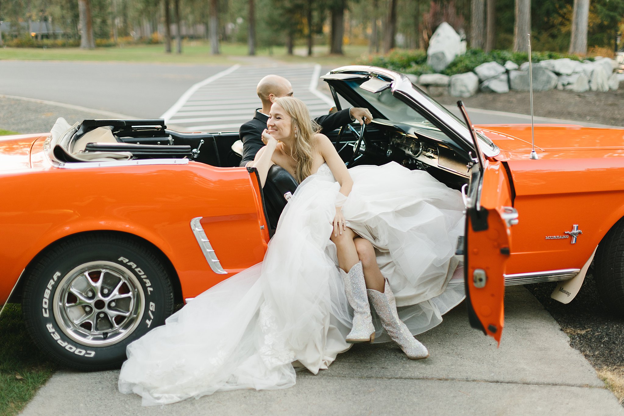 A bride smiles and relaxes in the front seat of a orange Ford Mustang in the driveway of the Kalispel Country Club Wedding venue