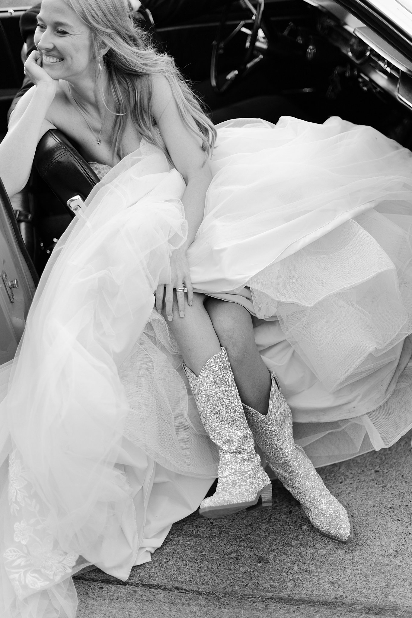 A bride in cowboy boots sits with legs hanging out a vintage car in black and white