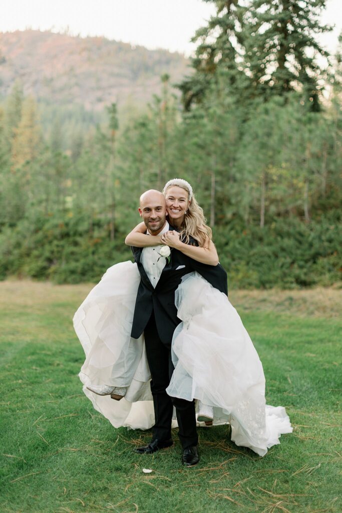 A bride hangs on the back of her groom in a field during their Kalispel Country Club Wedding