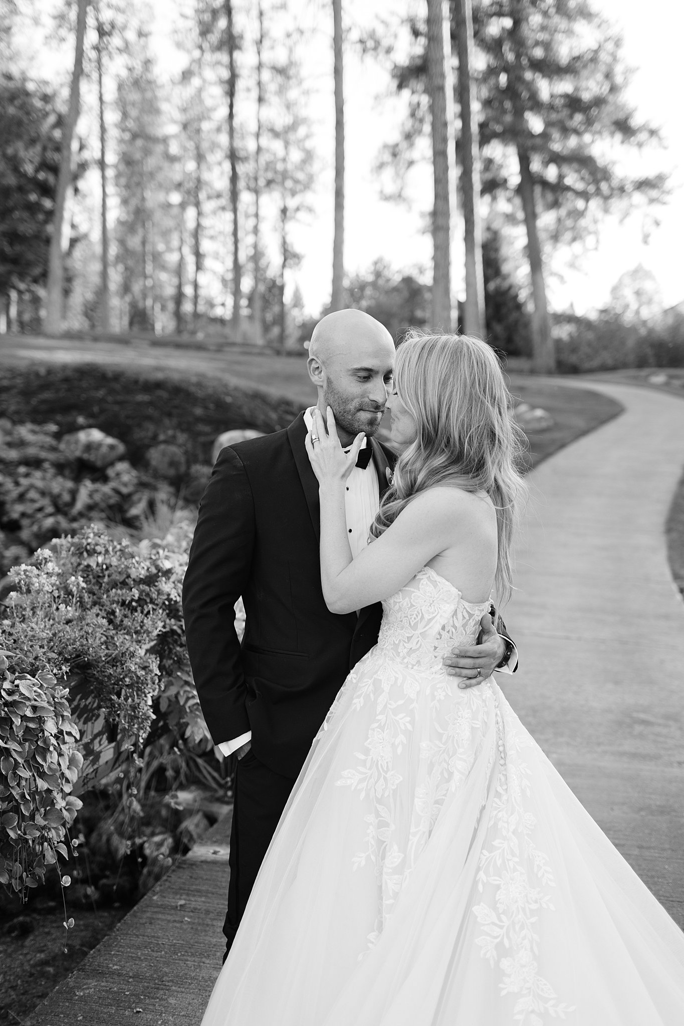 A bride and groom nuzzle while walking in a sidewalk path in black and white