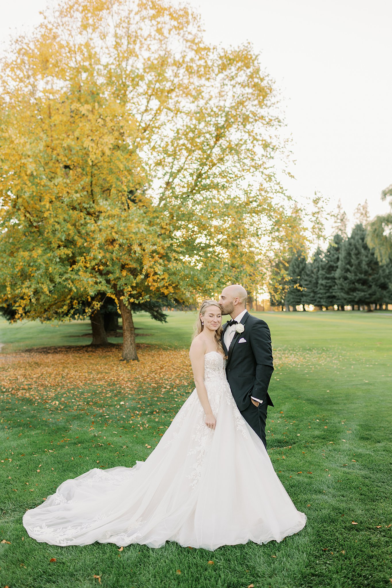 A groom in a black suit kisses the head of his smiling bride in a golf fairway at sunset at their Kalispel Country Club Wedding