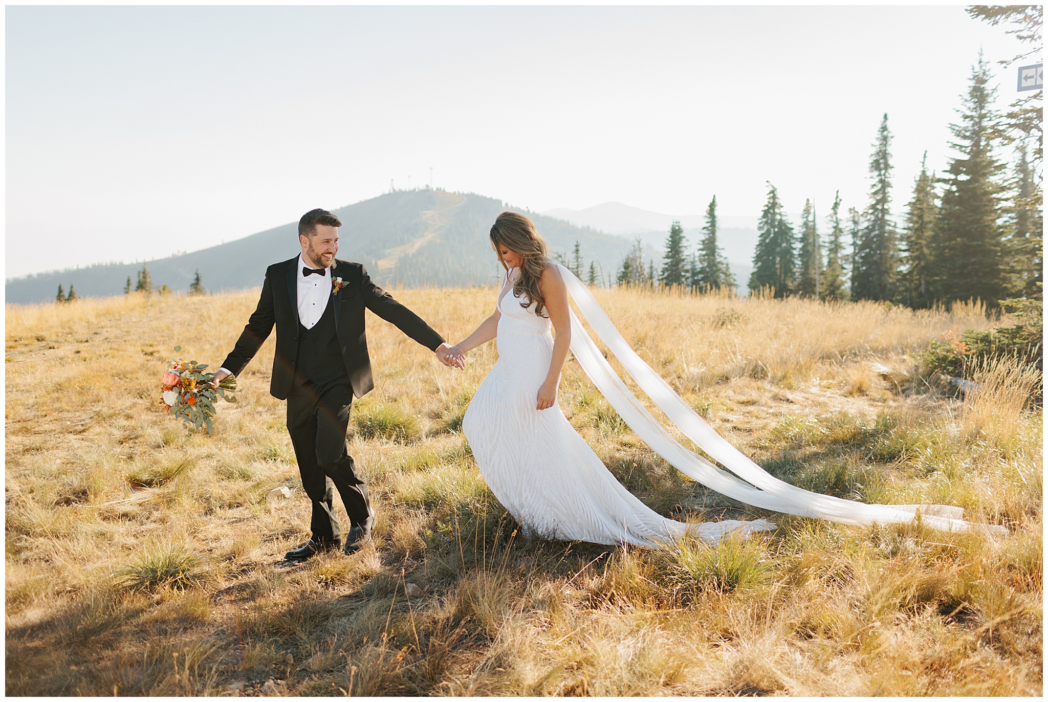 A groom in a black tuxedo walks his bride through a mountain pasture during their Schweitzer Mountain Wedding