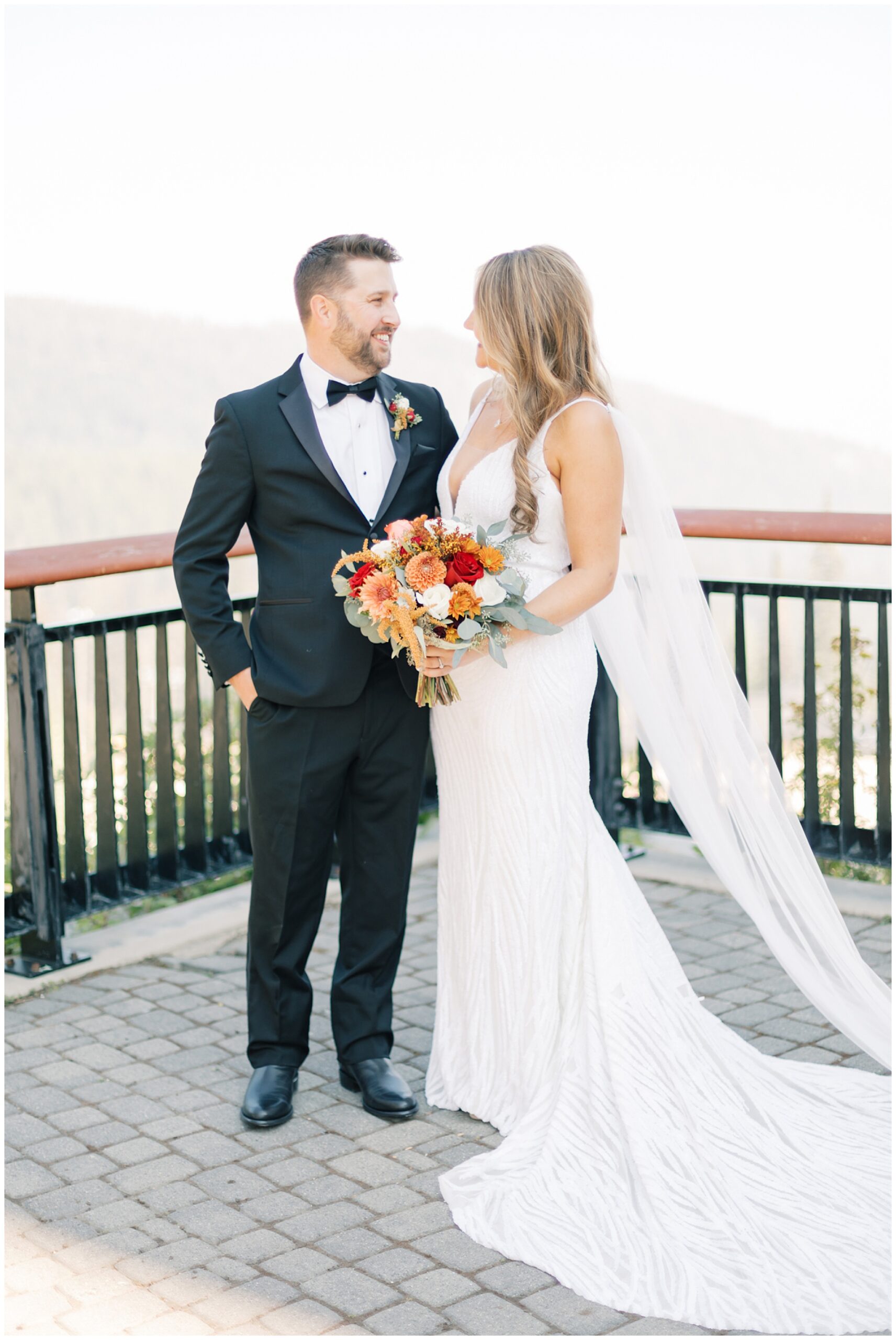 A bride and groom smile at each other while standing at a mountain overlook