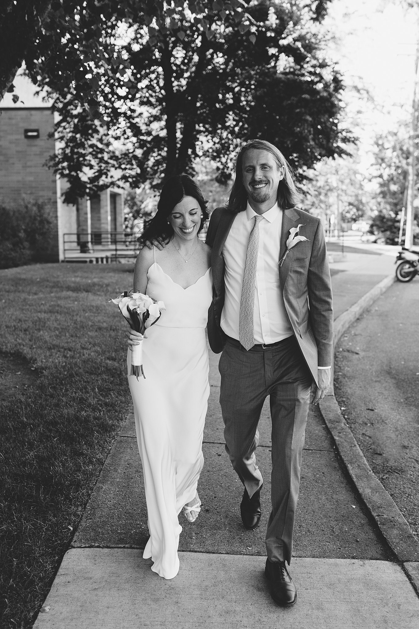 Newlyweds with arms around each other walk on a sidewalk smiling