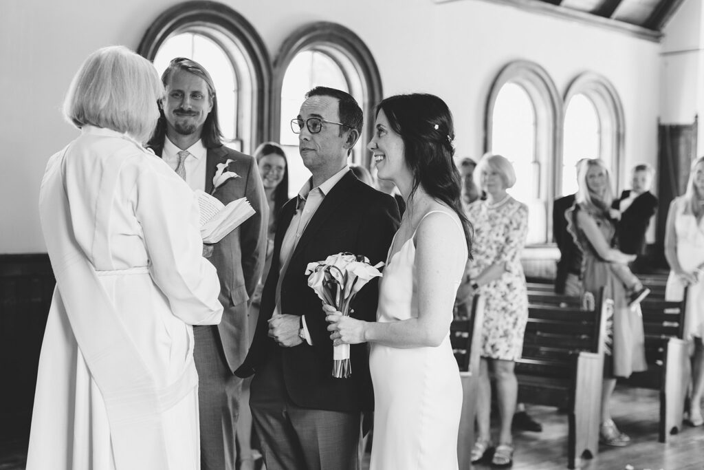 A bride smiles while being handed off by a relative to her groom at the altar